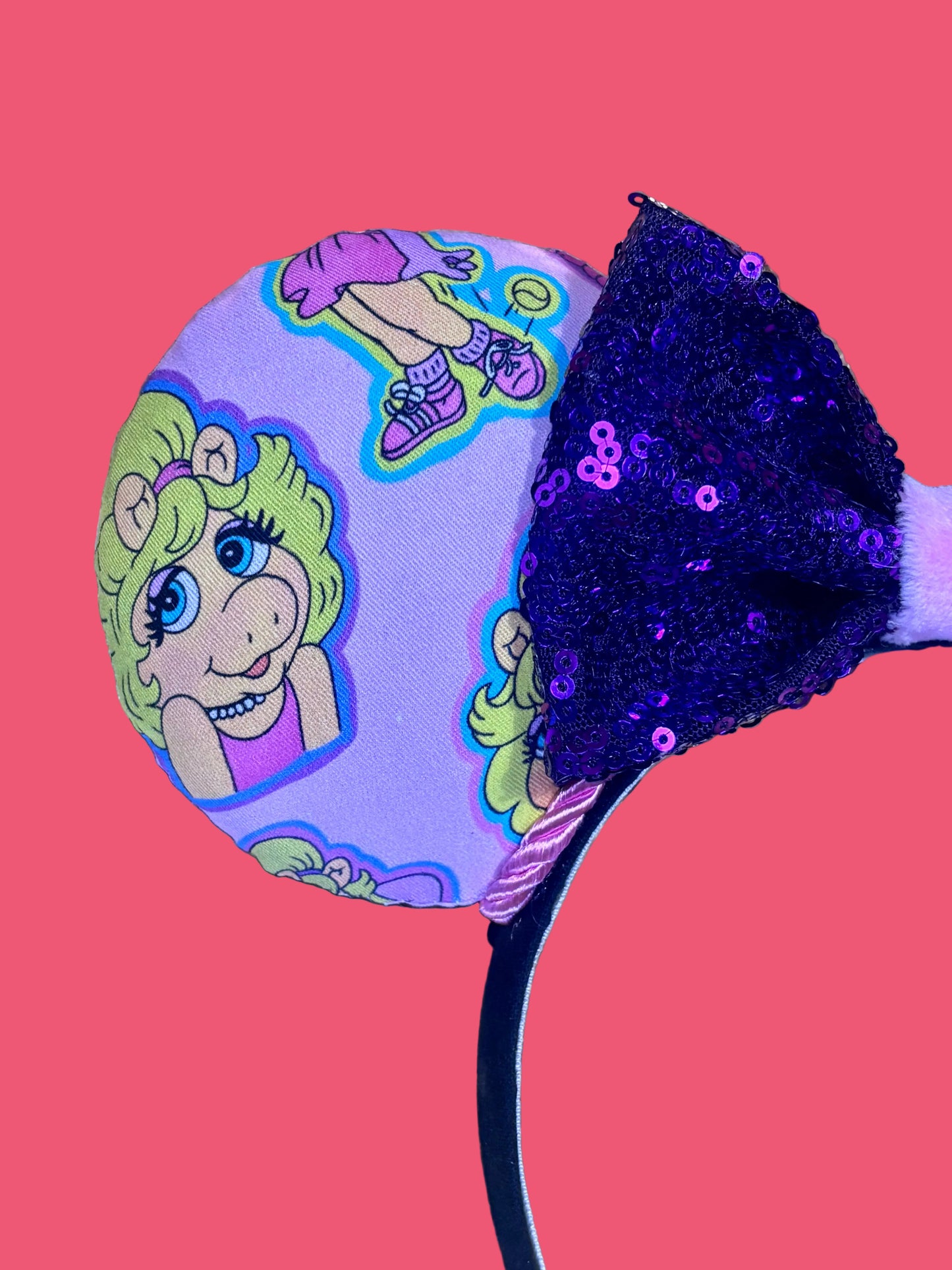 Handmade and ready to ship Piggy mouse ears. Miss Piggy fabric is on the front and the back. On a universal sized black poly fabric headband. With a purple sequin with pink soft middle on the bow. Close up of front left mouse ear.