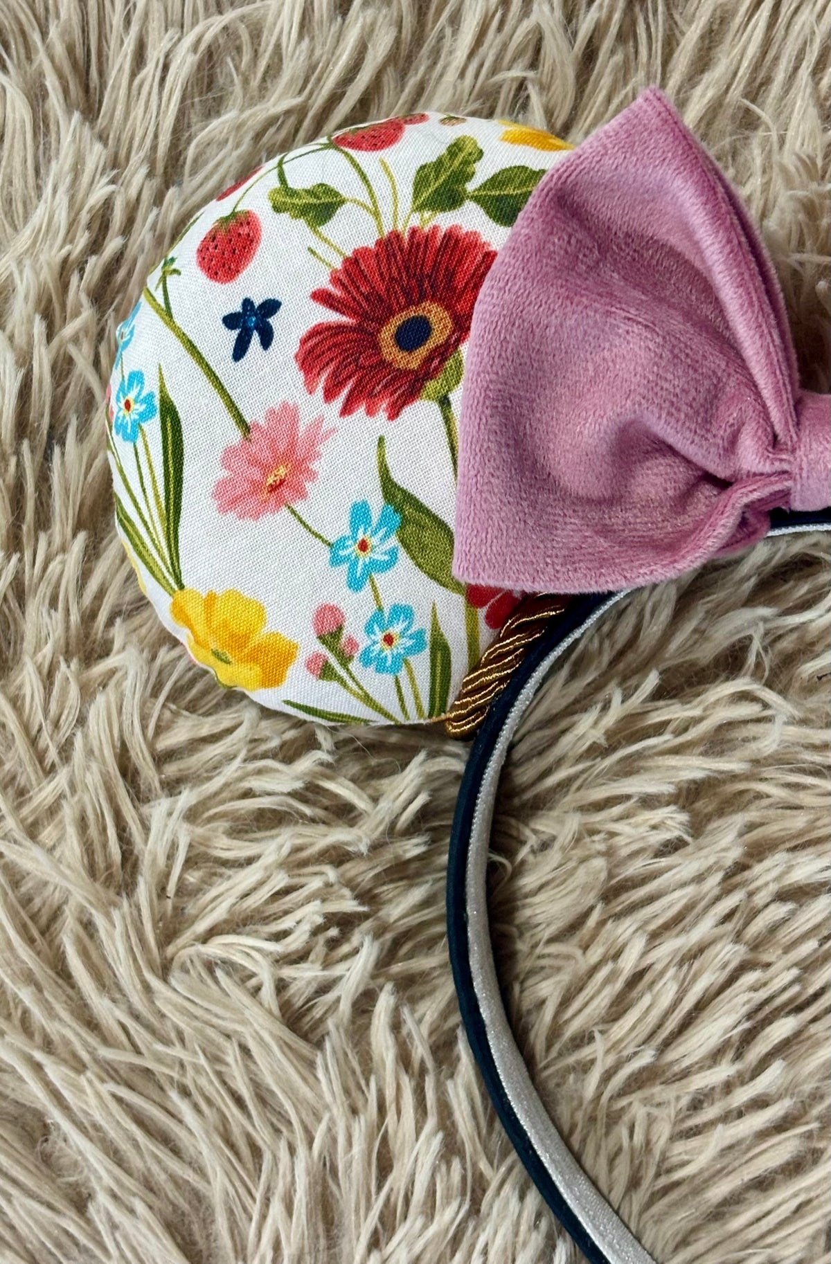 A close-up of a fabric-covered headband featuring a round floral ear and a large dusty pink bow, resting on a beige shag rug.