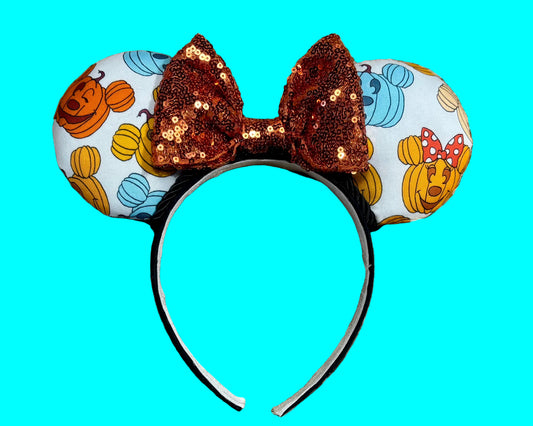 Pumpkin-themed ear headband featuring a light white background with orange, yellow, and blue pumpkins, a large sequined orange bow, and a black headband.