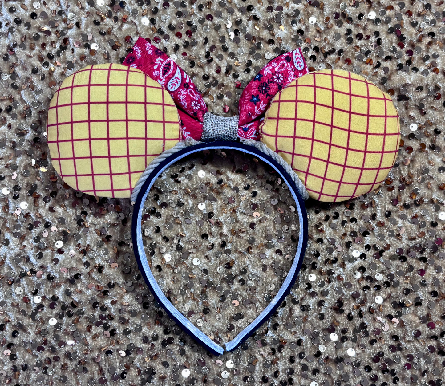 Handmade and ready to ship. Sheriff Woody Inspired mouse ears with the fabric on both the front and back that looks like his shirt. The fabric on the headband is black and is universal sized and fits most. The set has a bandana and cow print layered bow with brown trim along the inner ears to look like rope. Item is shown on a gold background.