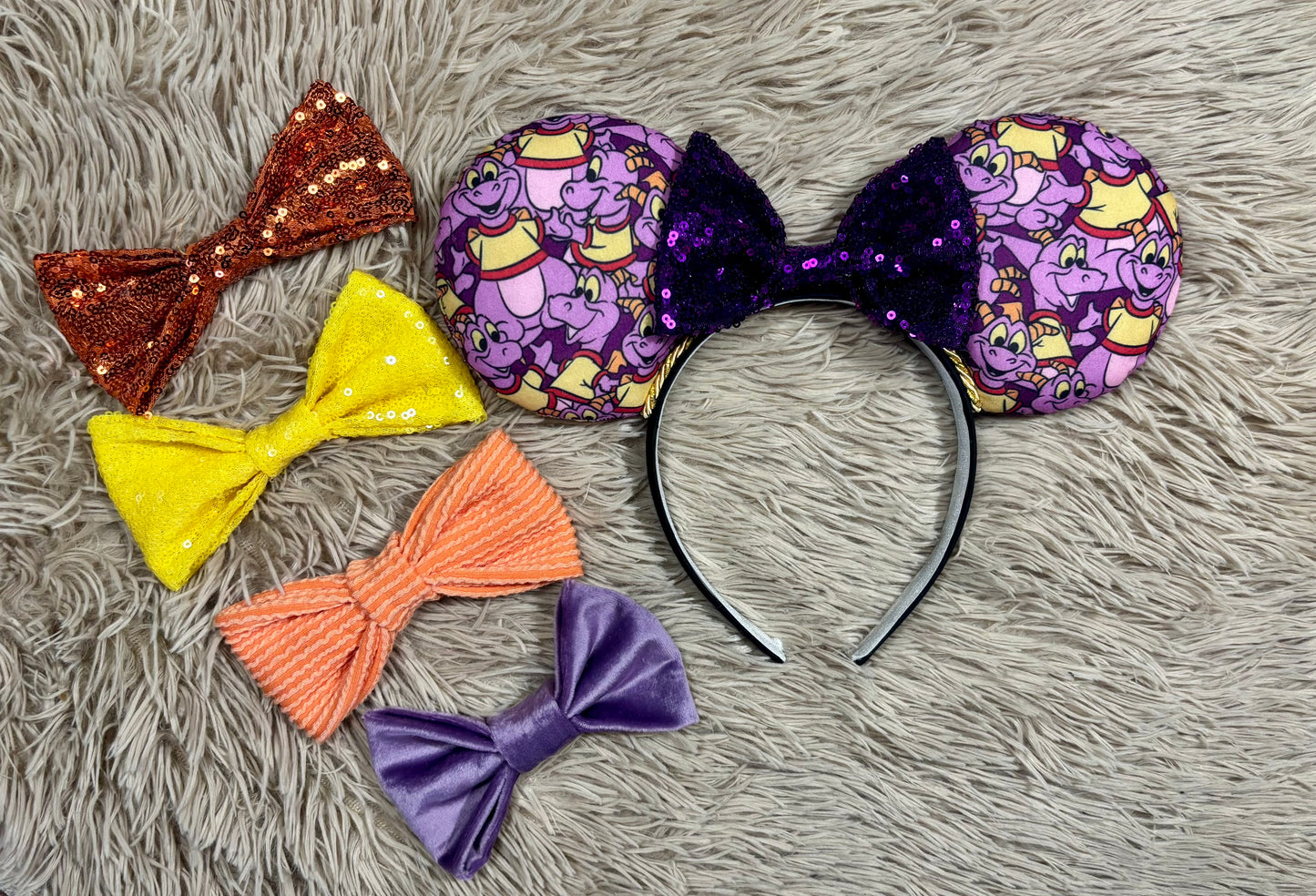 A pair of dragon style ears with a black headband, featuring a purple and yellow cartoon pattern on the ears and a dark purple sequined bow in the center. Shown with bow color options in case you don't want the purple bow. 