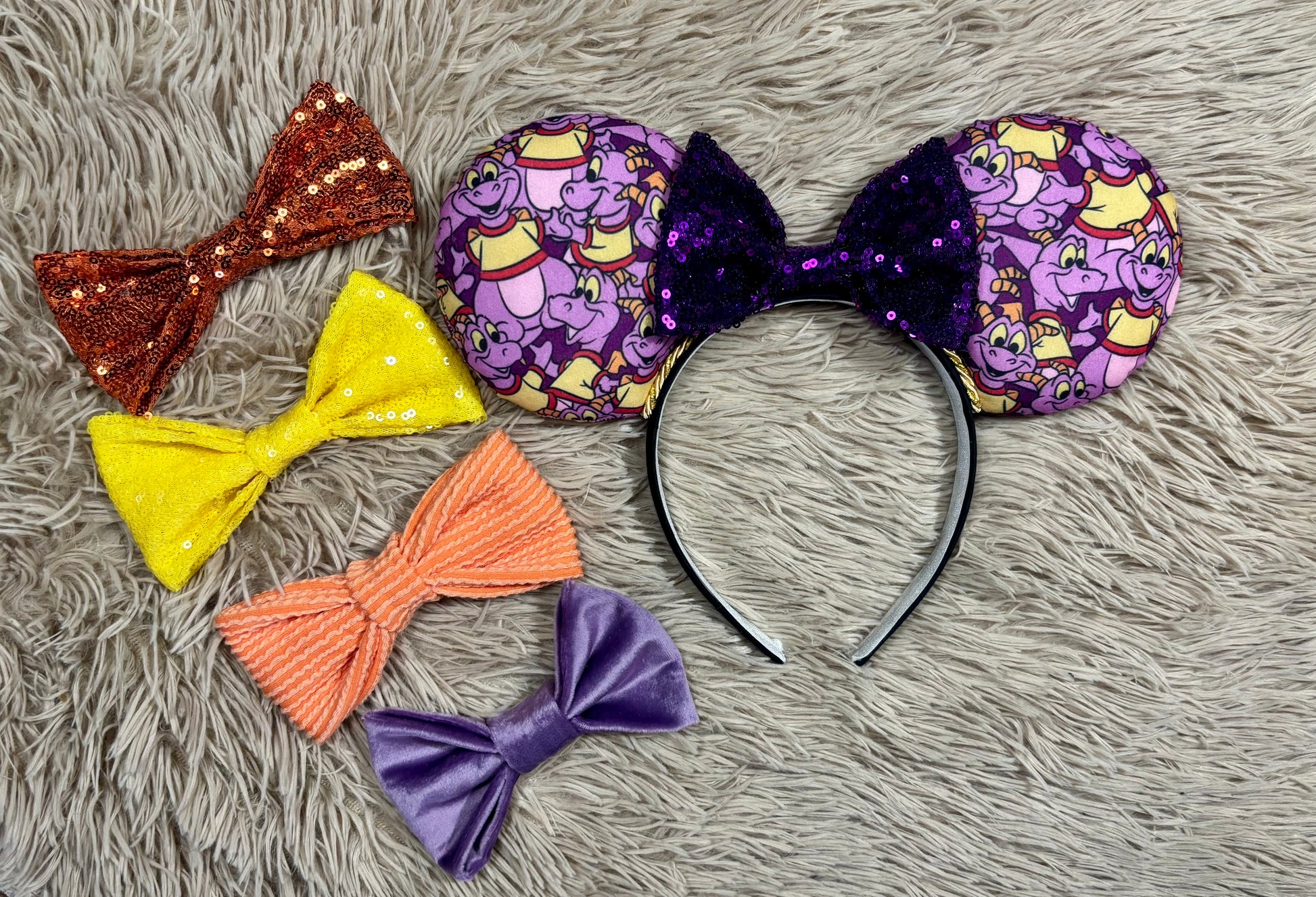 A pair of dragon style ears with a black headband, featuring a purple and yellow cartoon pattern on the ears and a dark purple sequined bow in the center. Shown with bow color options in case you don't want the purple bow. 