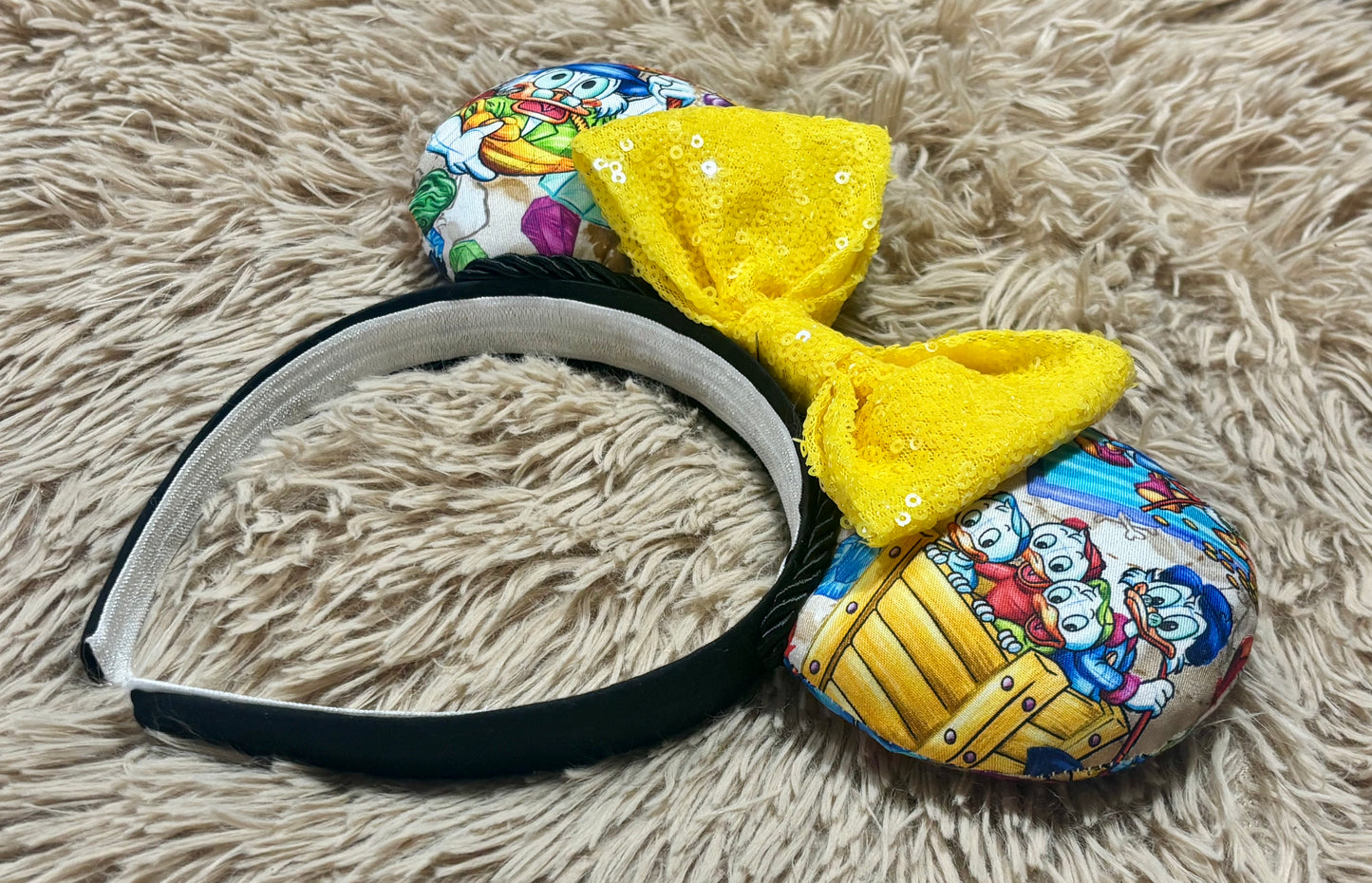 An ear headband featuring a large yellow sequined bow and cartoon ears with colorful printed graphics, displayed on a beige shag rug.