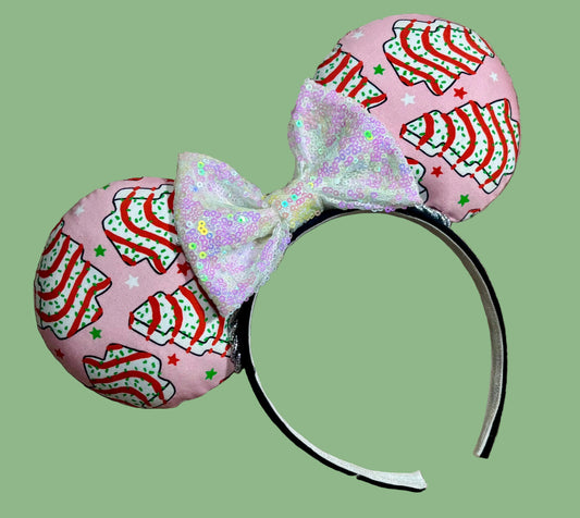 Pink ear headband with a glittery bow, featuring a red-and-white striped pattern with small green and white Christmas tree cakes, laid on a beige faux-fur surface.