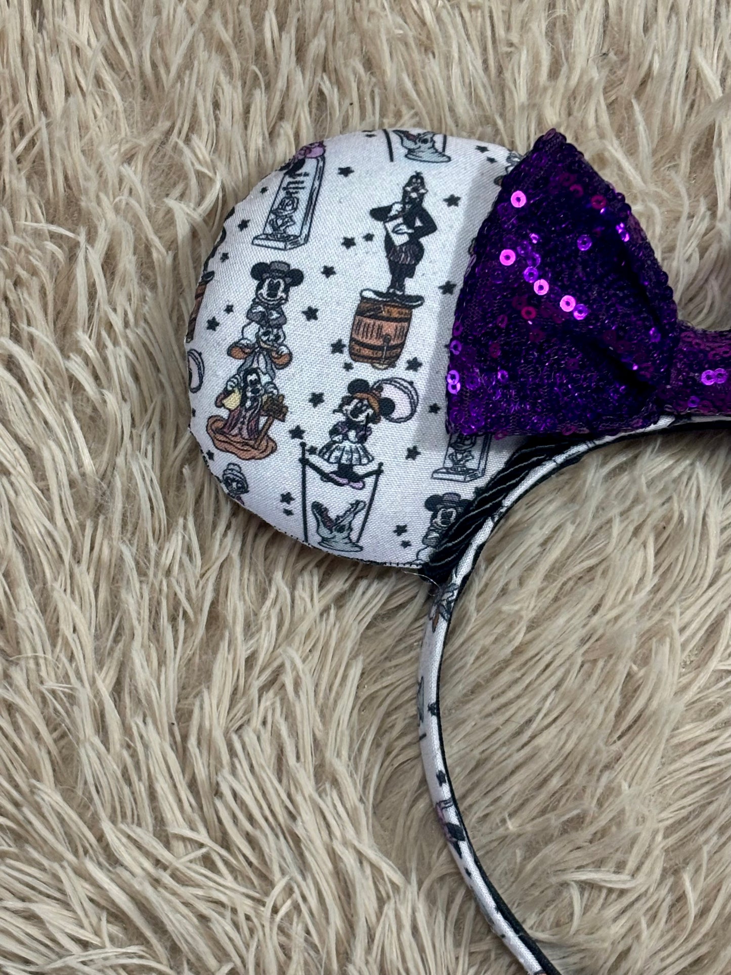 A close-up of a headband featuring a round white fabric ear with a black and gray cartoon character print and a large purple sequined bow on a beige faux-fur background.