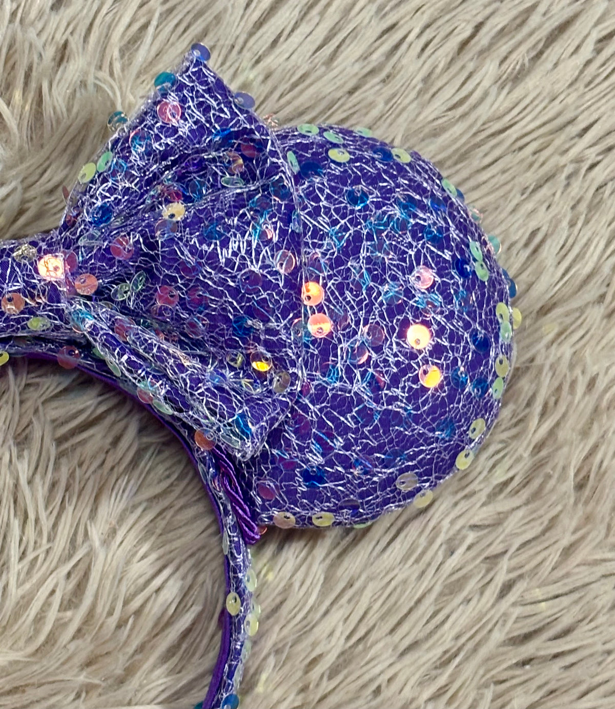 A purple sequined headband with large round ears and a bow, laid flat on a beige faux fur surface. Close up of the mouse ear.
