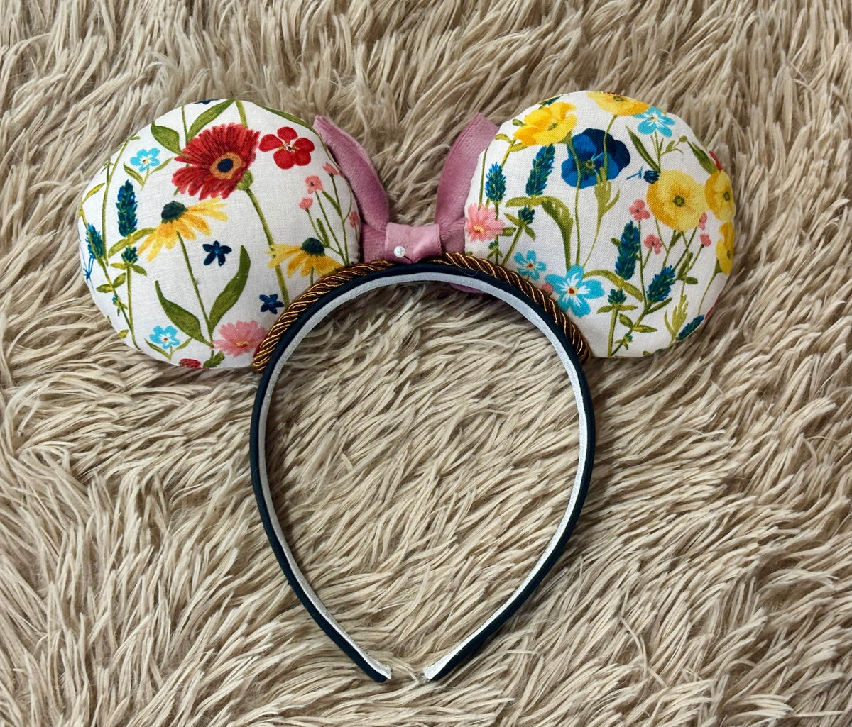 An ear headband accessory with a colorful wildflower print on a beige faux-fur background. The fabric ears feature red, yellow, blue, pink, and green flowers on a cream background, attached to a black headband with a pink bow. Back side view of the ears.