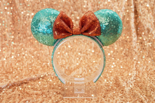 Glittery green and orange mouse ears inspired for the showgirls. Rhinestone trim is on the ears as well. Shown on a clear stand against a glittery gold background