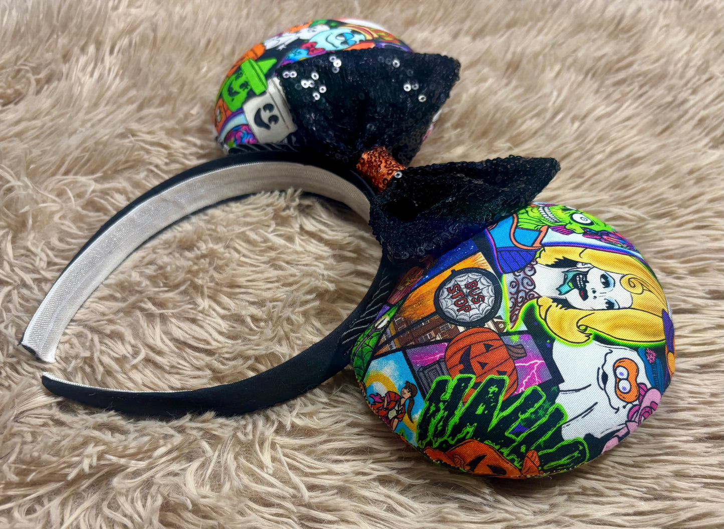 An ear headband accessory featuring a colorful Halloween-themed print on the outer ears and a black glitter bow in the center with black twisted trim along the inner ears. Side view of the set.