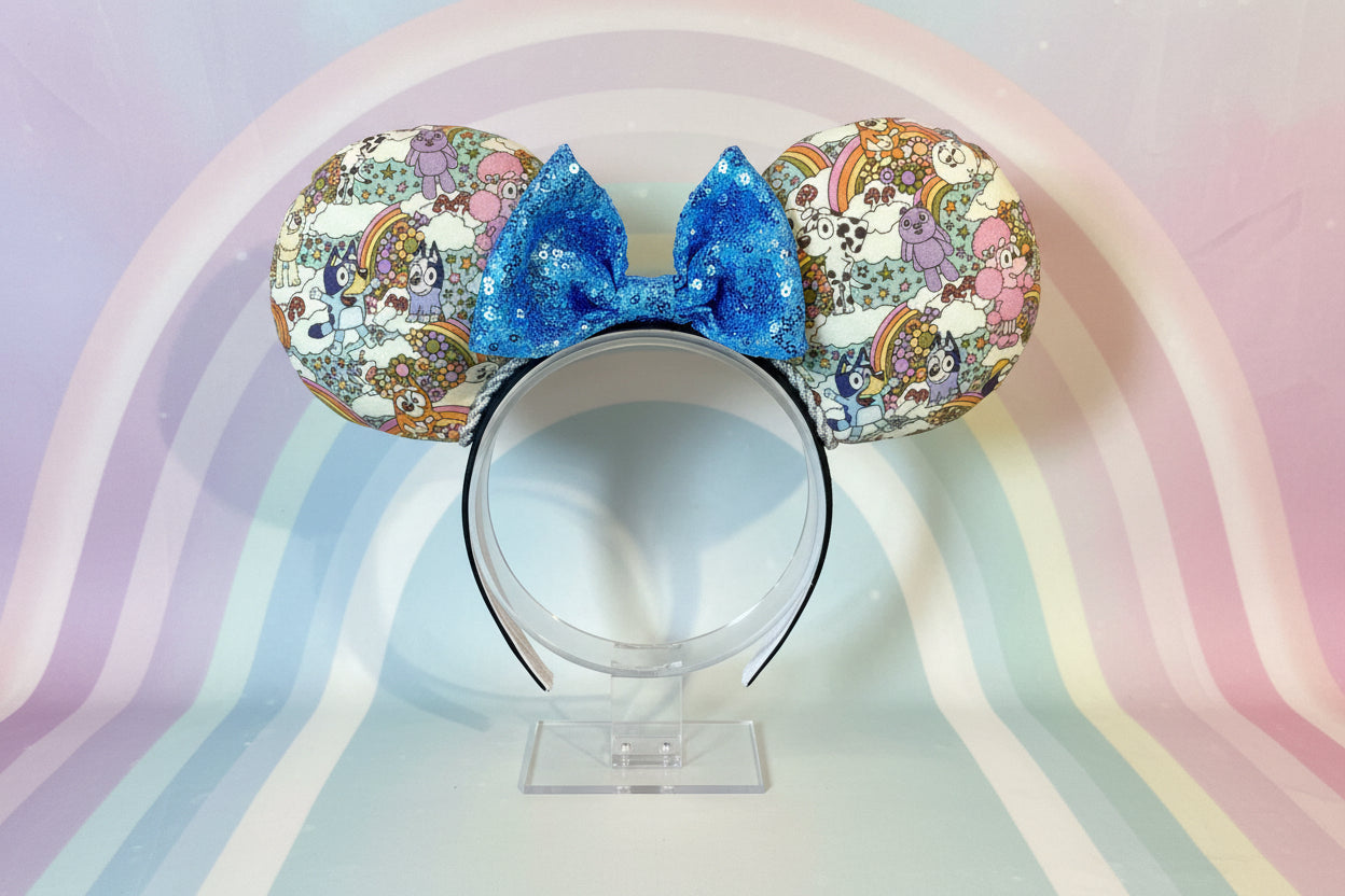 Colorful Bluey inspired fabric is on the Mouse ears with a light blue sequin bow on a black headband. Shown on a stand against a pastel rainbow background