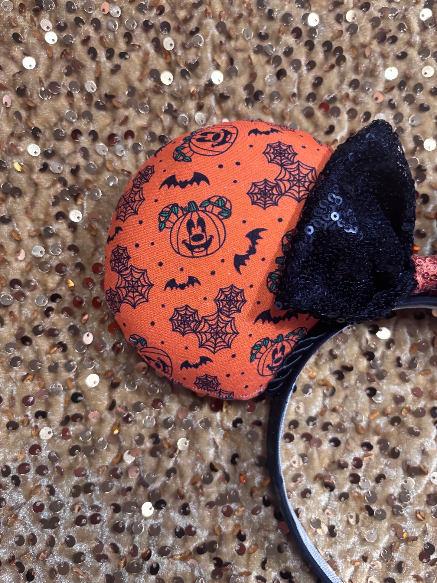 Halloween mouse ears with the fabric on both the front and back. It's orange with bats, mouse shaped pumpkins and spider webs. The headband is black universal sized and fits most. The set has a black with orange middle sequin bow. Item is shown on a gold sequin background.