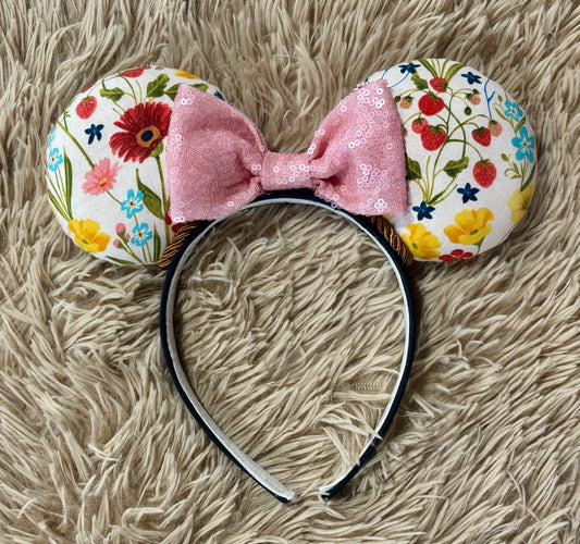 A handmade ear headband featuring a white fabric with a colorful floral print and a light pink sequined bow, displayed on a beige shag rug.