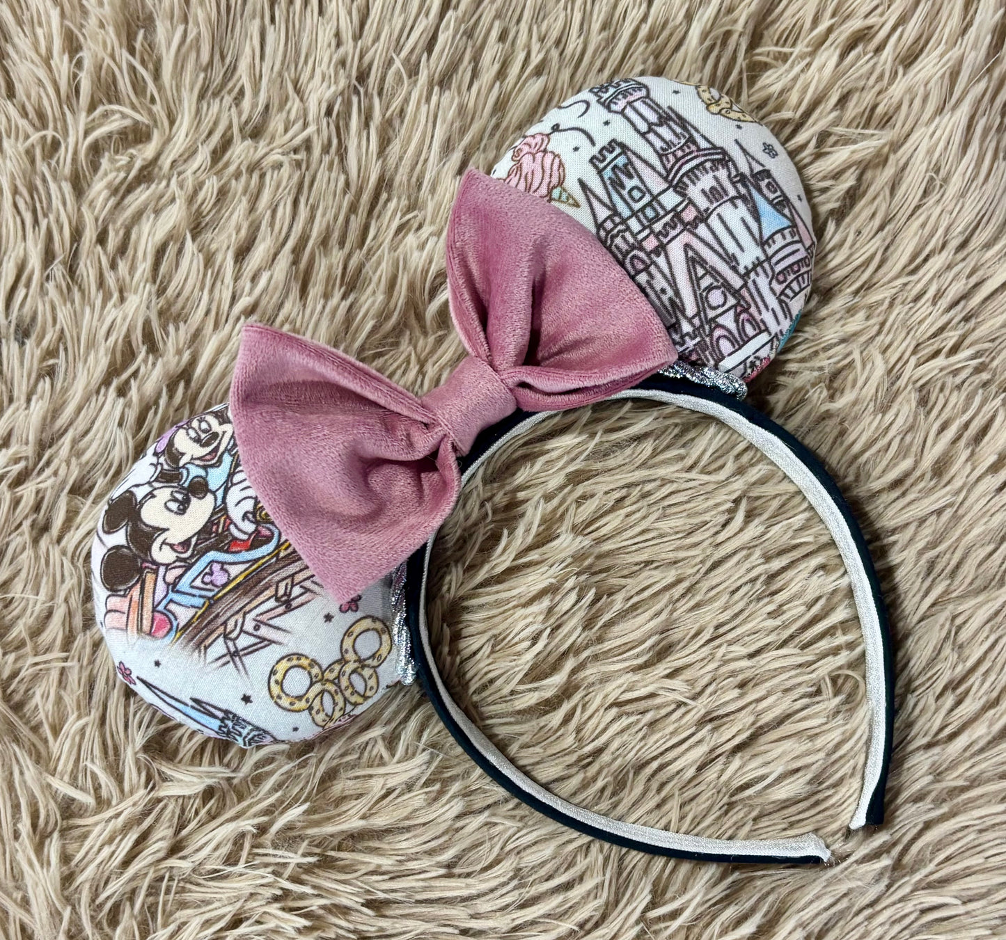 A pair of handmade ear headbands with a large dusty pink velvet bow, featuring printed park themed patterns on the ears and resting on a beige faux fur surface.