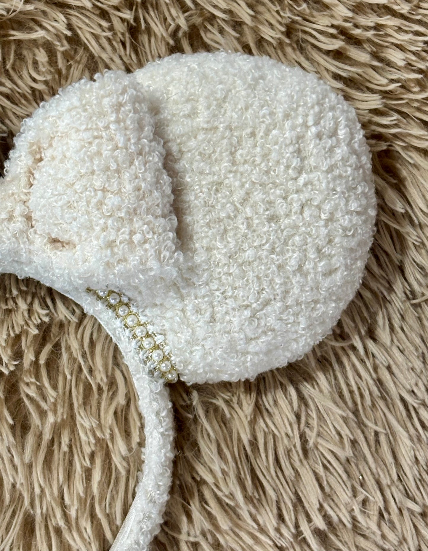 A close-up of a textured cream-colored headband with a small decorative gold embellishment, laid flat on a beige shaggy rug.