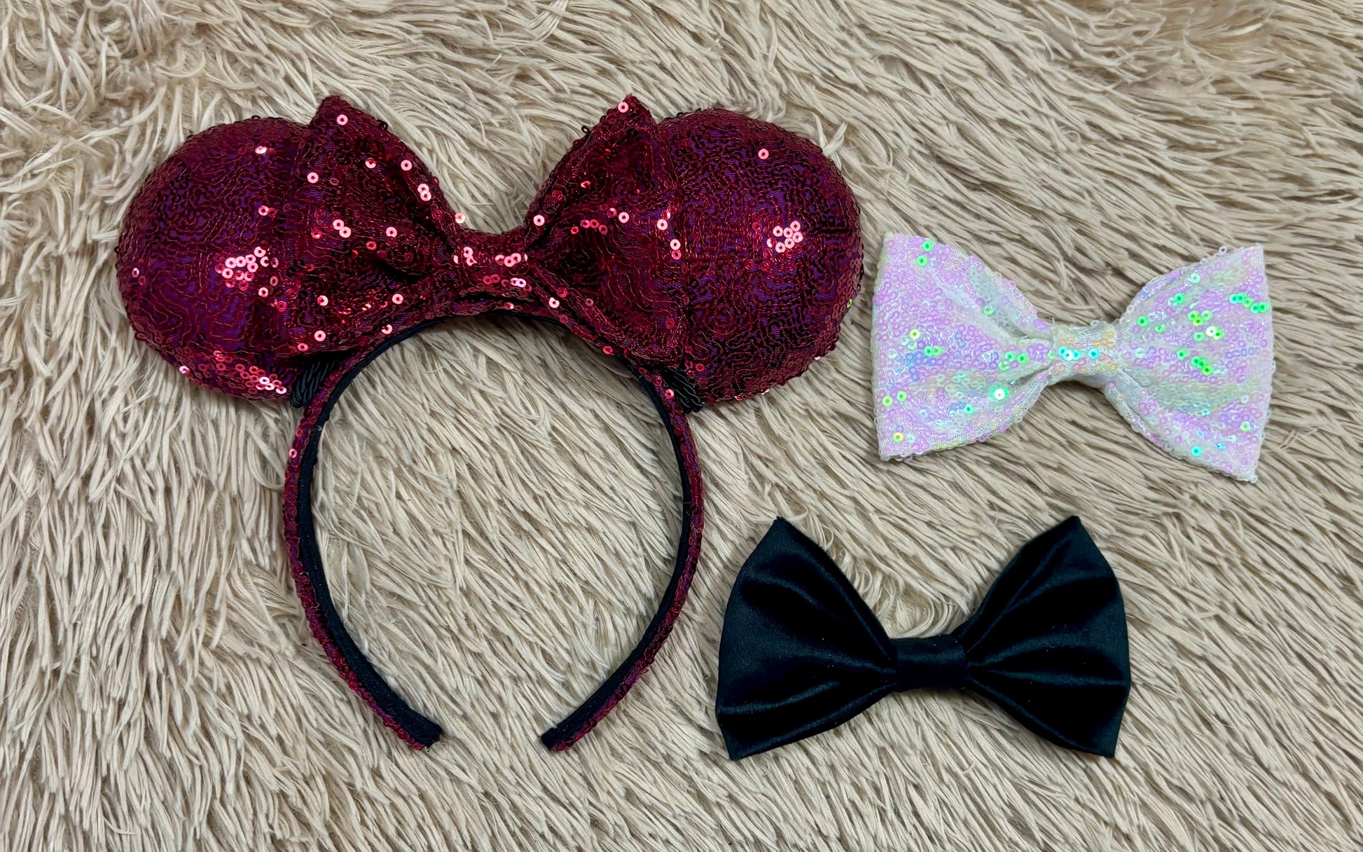 Three sequined bow headbands laid flat on a beige faux-fur surface: one large burgundy sequin mouse-ear style headband, one iridescent white sequin bow, and one black velvet bow.
