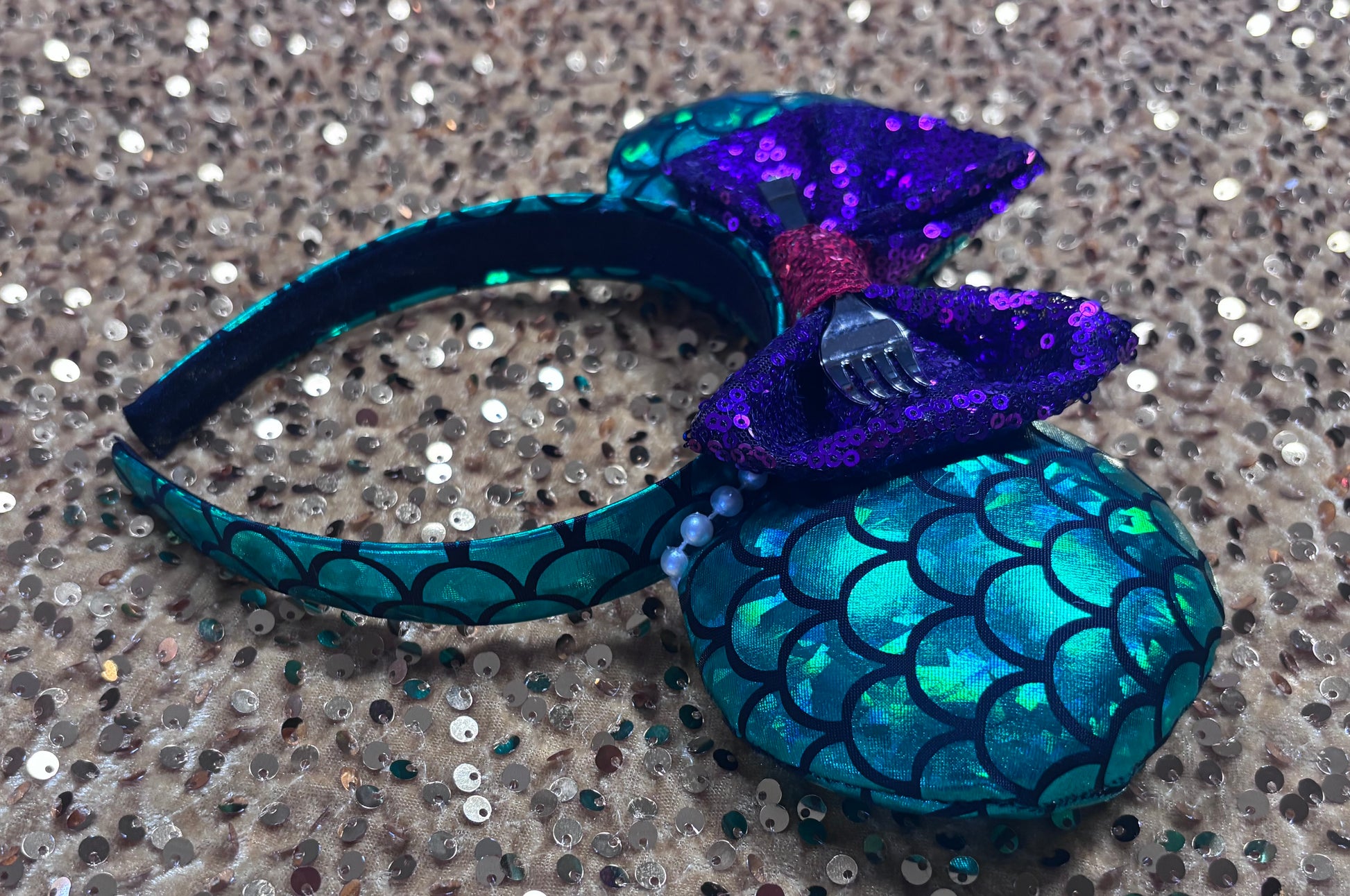 Side view of headband. Mermaid-themed headband with teal scales and a purple sequin bow with a red middle and a dinglehopper middle piece. Pearl garland trim along inner ears m on a matching mermaid scales headband.