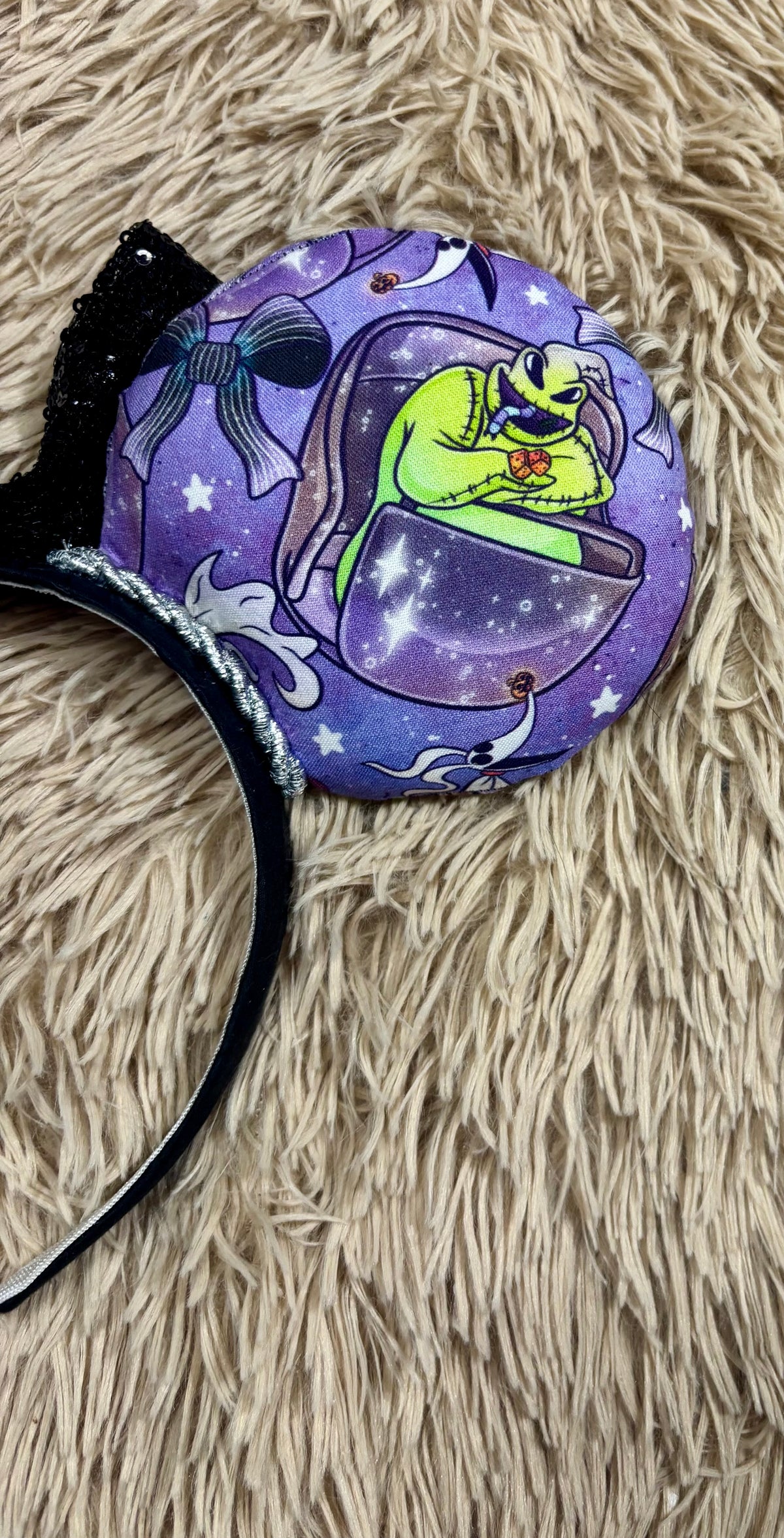 An ear headband accessory with a black sequined bow, featuring a purple HM and NBC themed print with a cartoon character in a buggy.