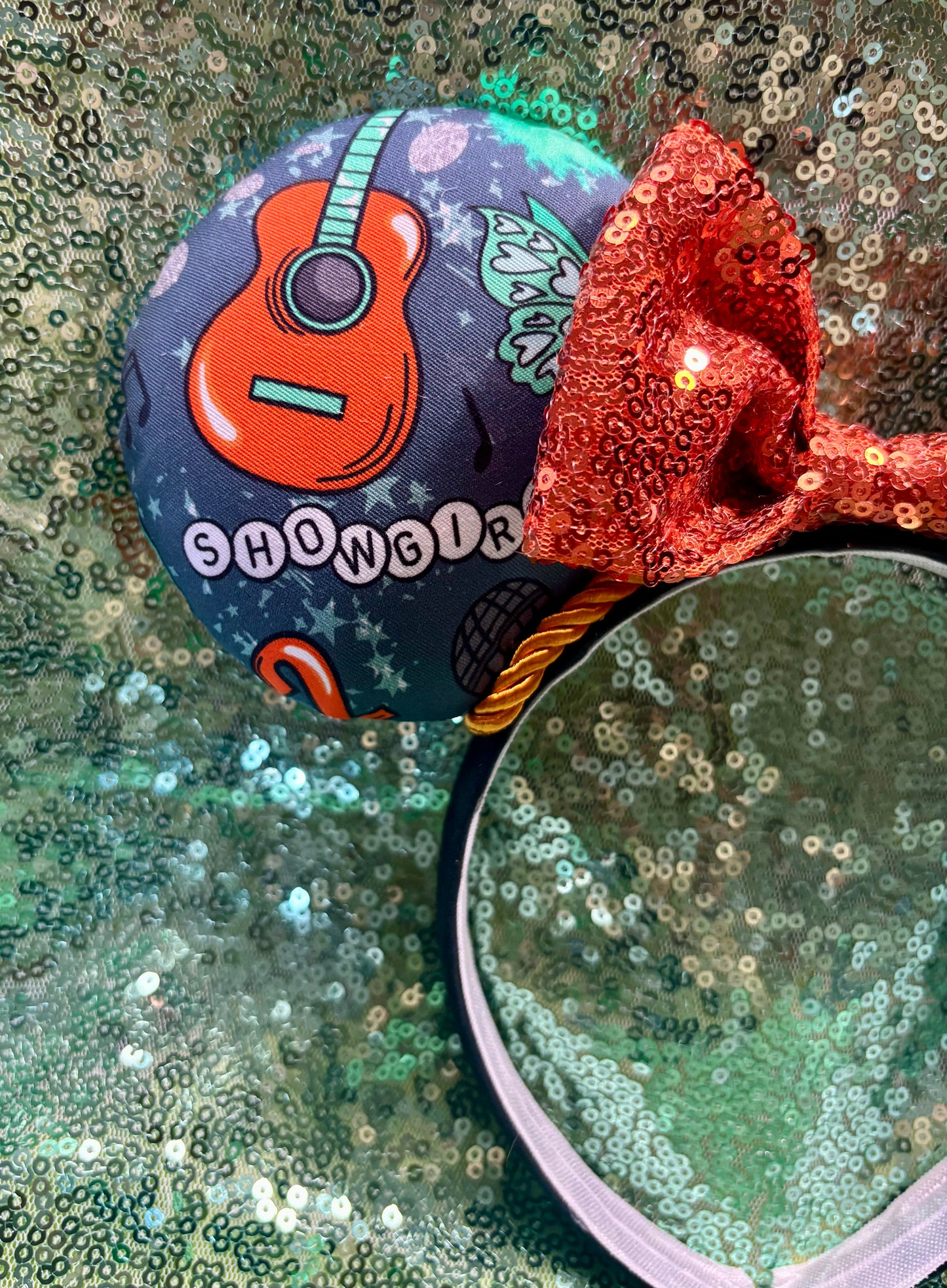 Close up of mouse ear with a guitar and the words showgirl. Handmade and ready to ship Actually Romantic Mouse ears with an orange sequin bow, orange twisted trim and on a black universal sized headband.