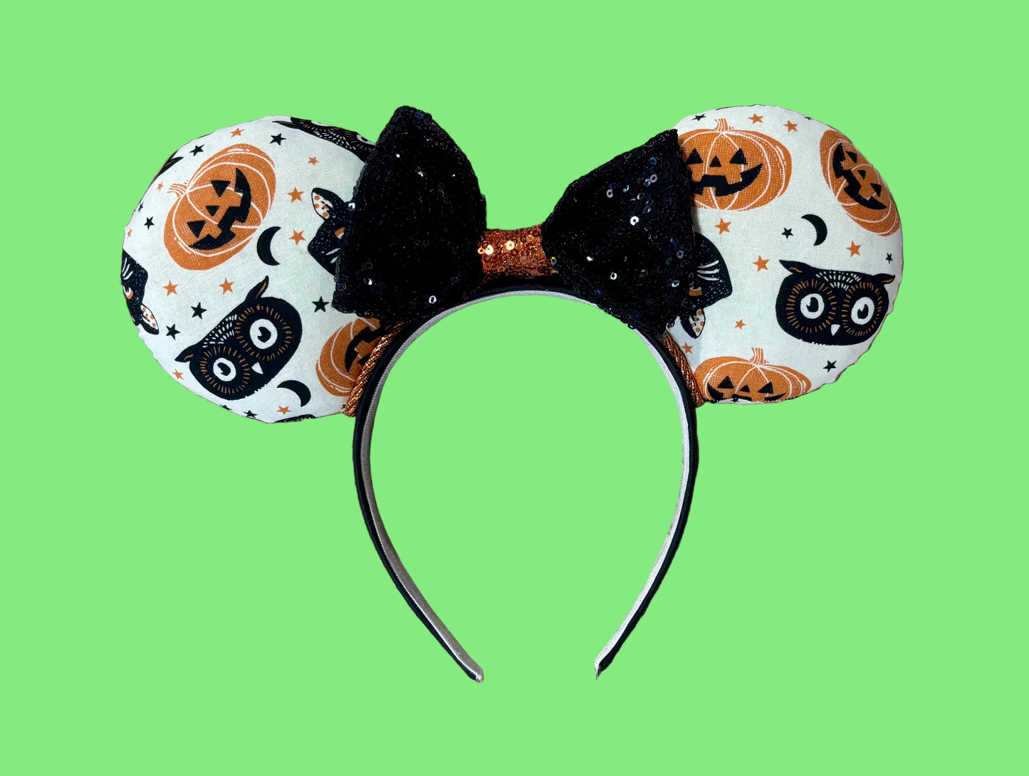 A Halloween-themed ear headband featuring a white fabric print with black cats, orange pumpkins, and stars, accented by a large black sequined bow with an orange glittery center.