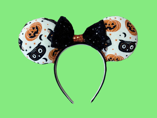 A Halloween-themed ear headband featuring a white fabric print with black cats, orange pumpkins, and stars, accented by a large black sequined bow with an orange glittery center.
