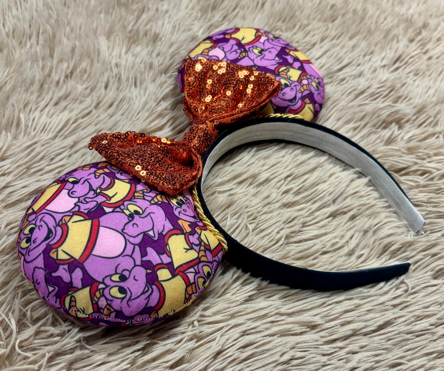 A pair of dragon style ears with a black headband, featuring a purple and yellow cartoon pattern on the ears and a dark purple sequined bow in the center