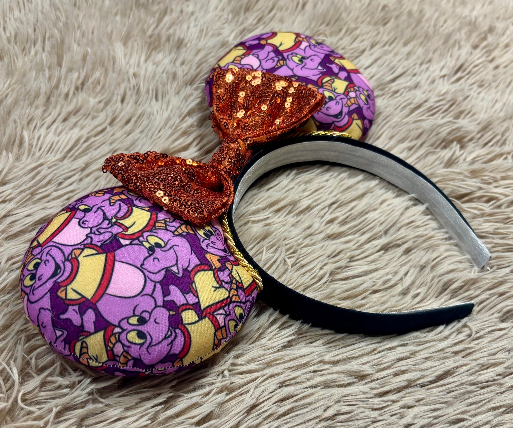 A pair of dragon style ears with a black headband, featuring a purple and yellow cartoon pattern on the ears and a dark purple sequined bow in the center