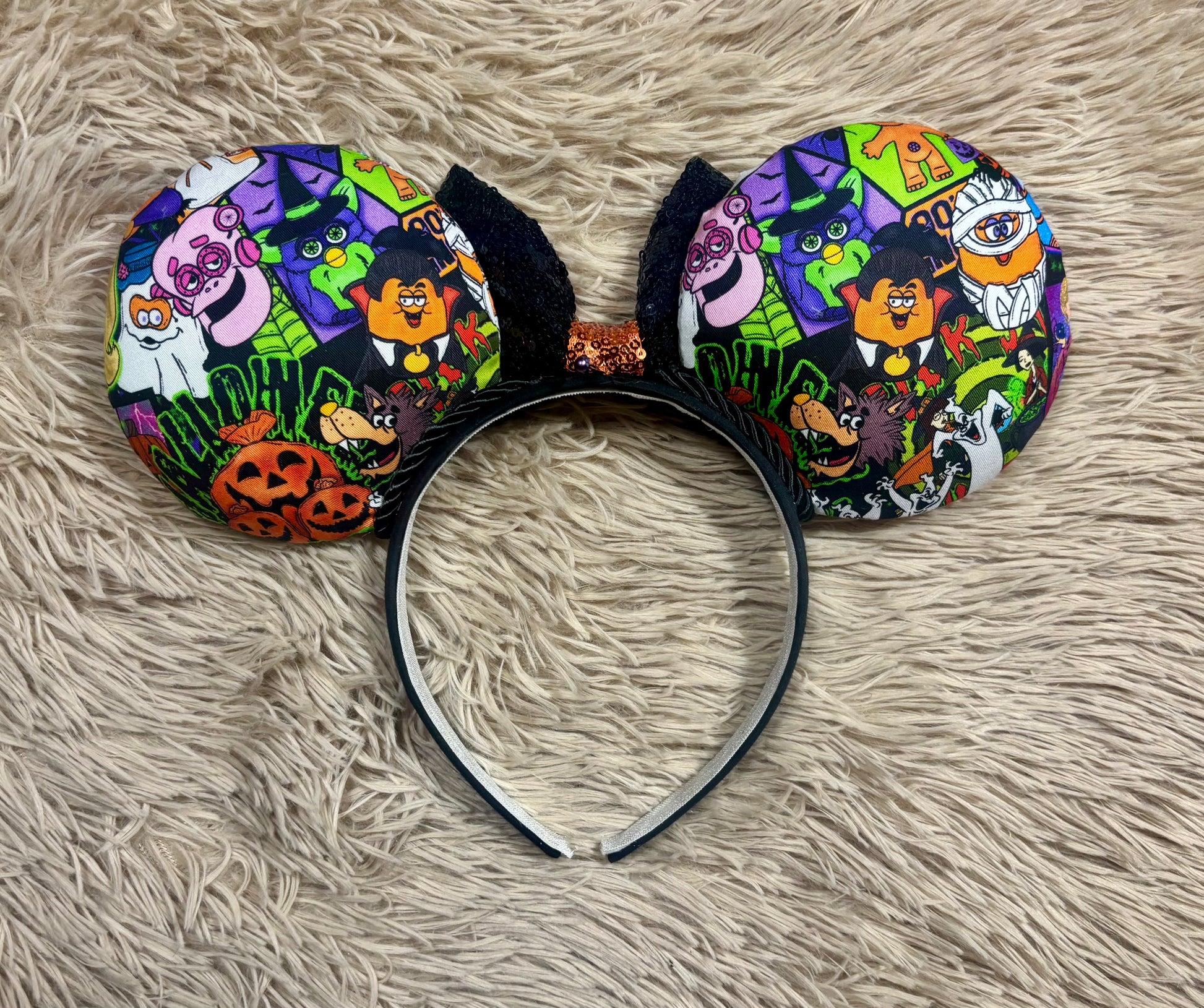 The back side view of the Halloween-themed mouse ears headband featuring a colorful cartoon print of pumpkins, ghosts, witches, and spooky characters on a black headband, laid flat on a beige faux fur background.