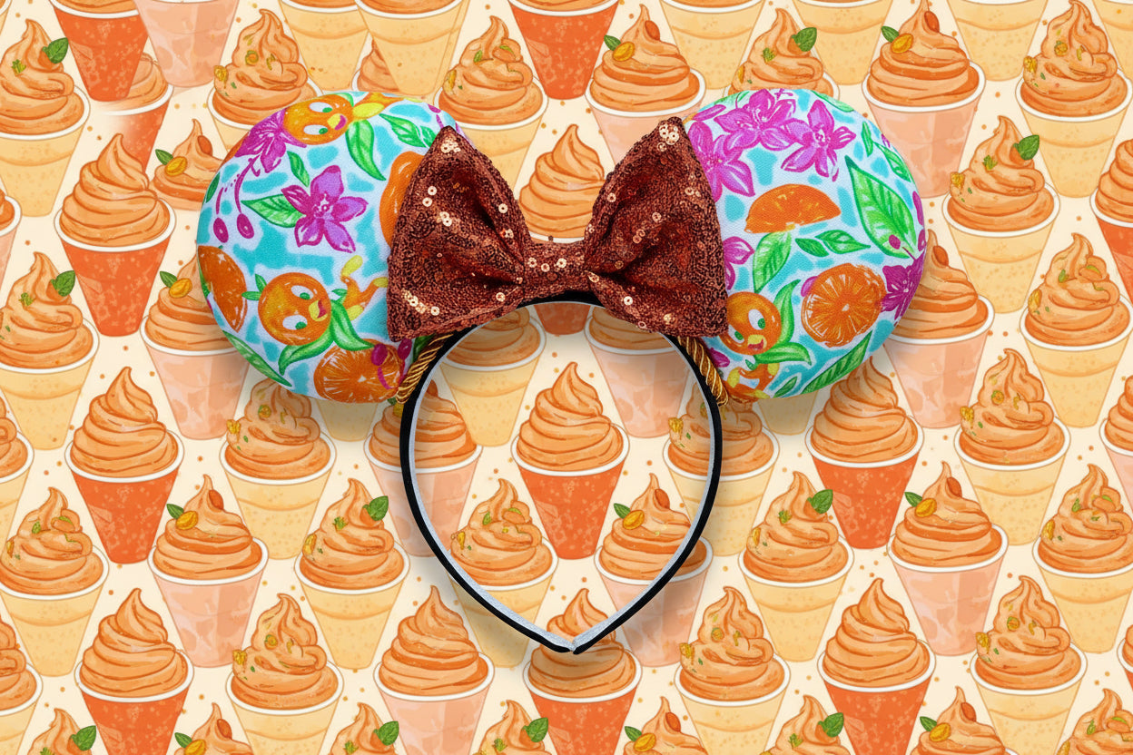 Colorful floral orange bird inspired fabric on the mouse ears with an orange sequin bow on a black headband. Shown on a glittery gold background 