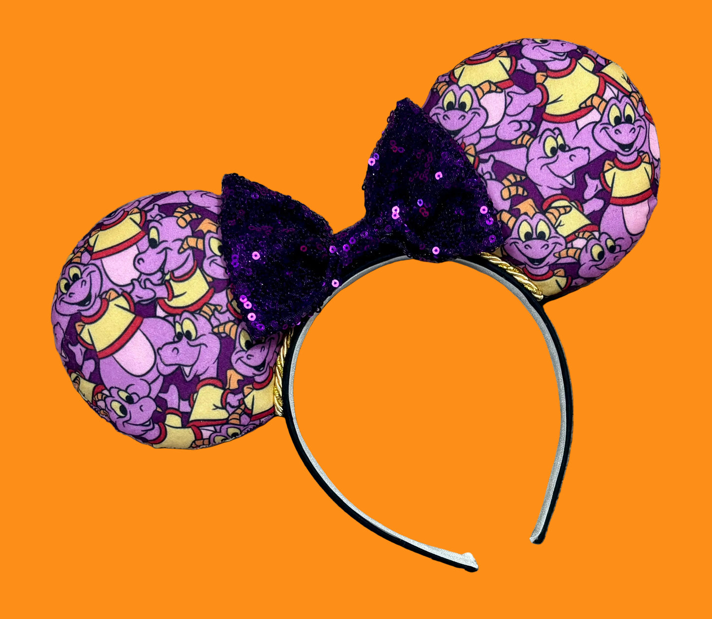 A pair of dragon style ears with a black headband, featuring a purple and yellow cartoon pattern on the ears and a dark purple sequined bow in the center.