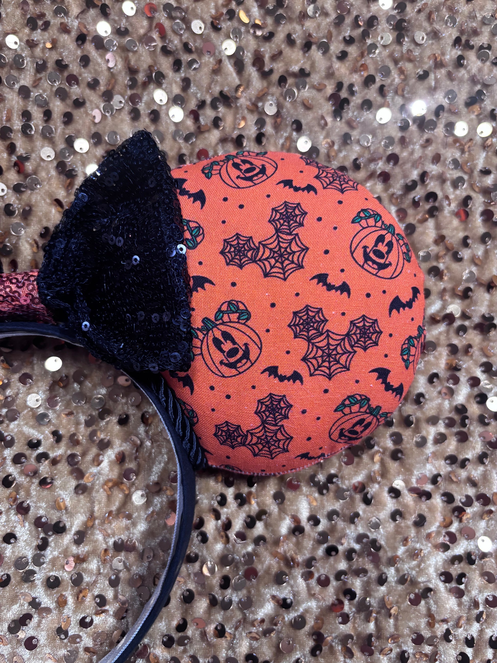 Halloween mouse ears with the fabric on both the front and back. It's orange with bats, mouse shaped pumpkins and spider webs. The headband is black universal sized and fits most. The set has a black with orange middle sequin bow. Item is shown on a gold sequin background.