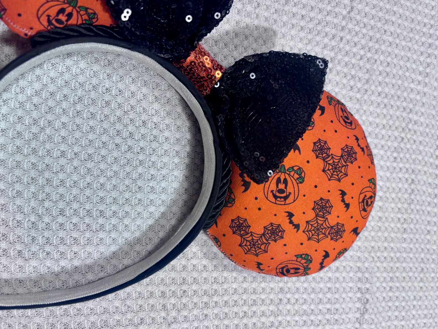 Halloween mouse ears with the fabric on both the front and back. It's orange with bats, mouse shaped pumpkins and spider webs. The headband is black universal sized and fits most. The set has a black with orange middle sequin bow. Item is shown on a white background. Close up of designs on fabric