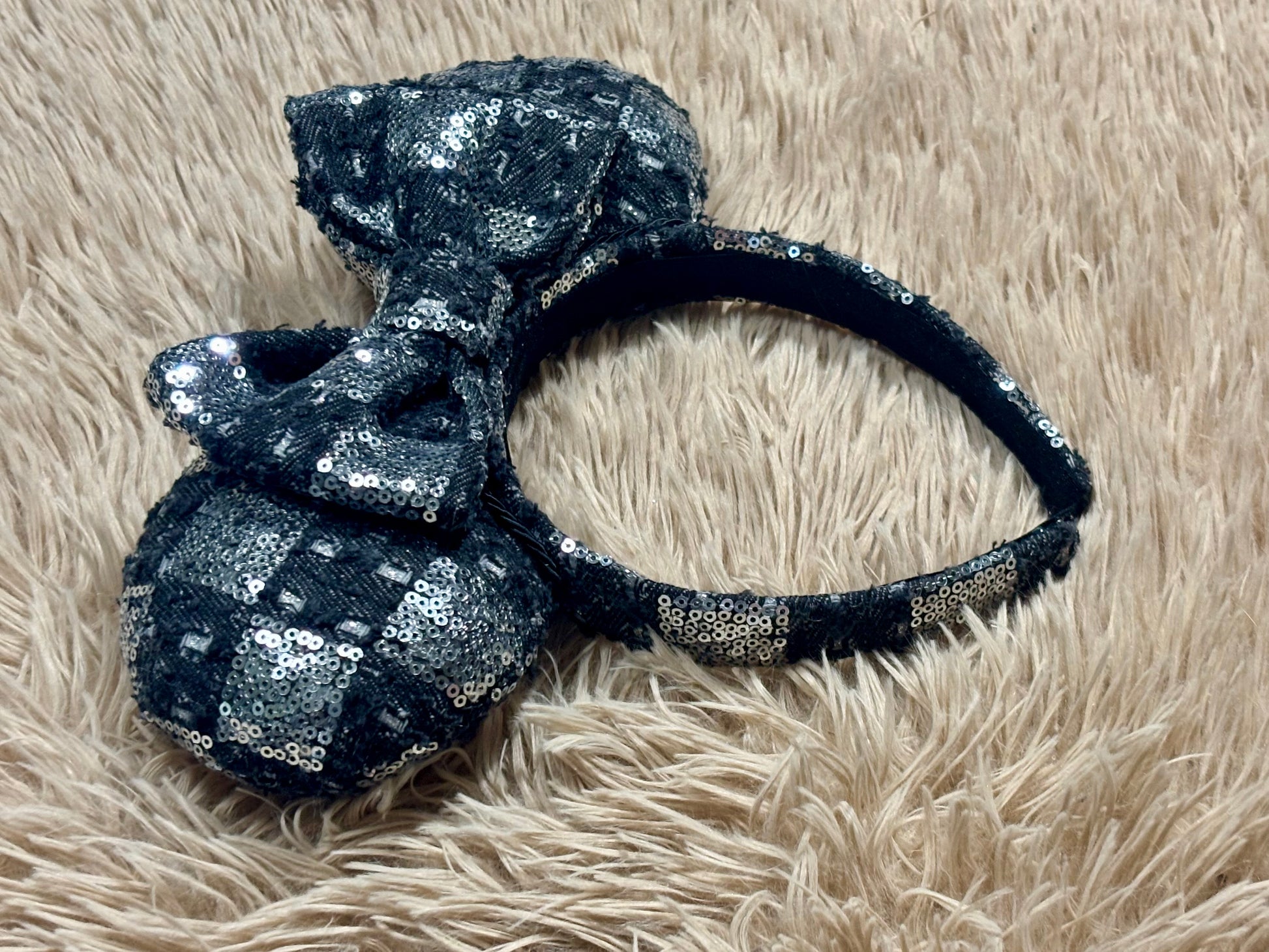 A black and silver checkered sequined headband with a large matching bow, displayed on a beige shag rug.