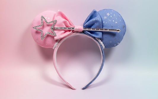 A pink and blue fabric ear headband with a silver rhinestone star wand accessory, set against a pastel pink and blue gradient background.