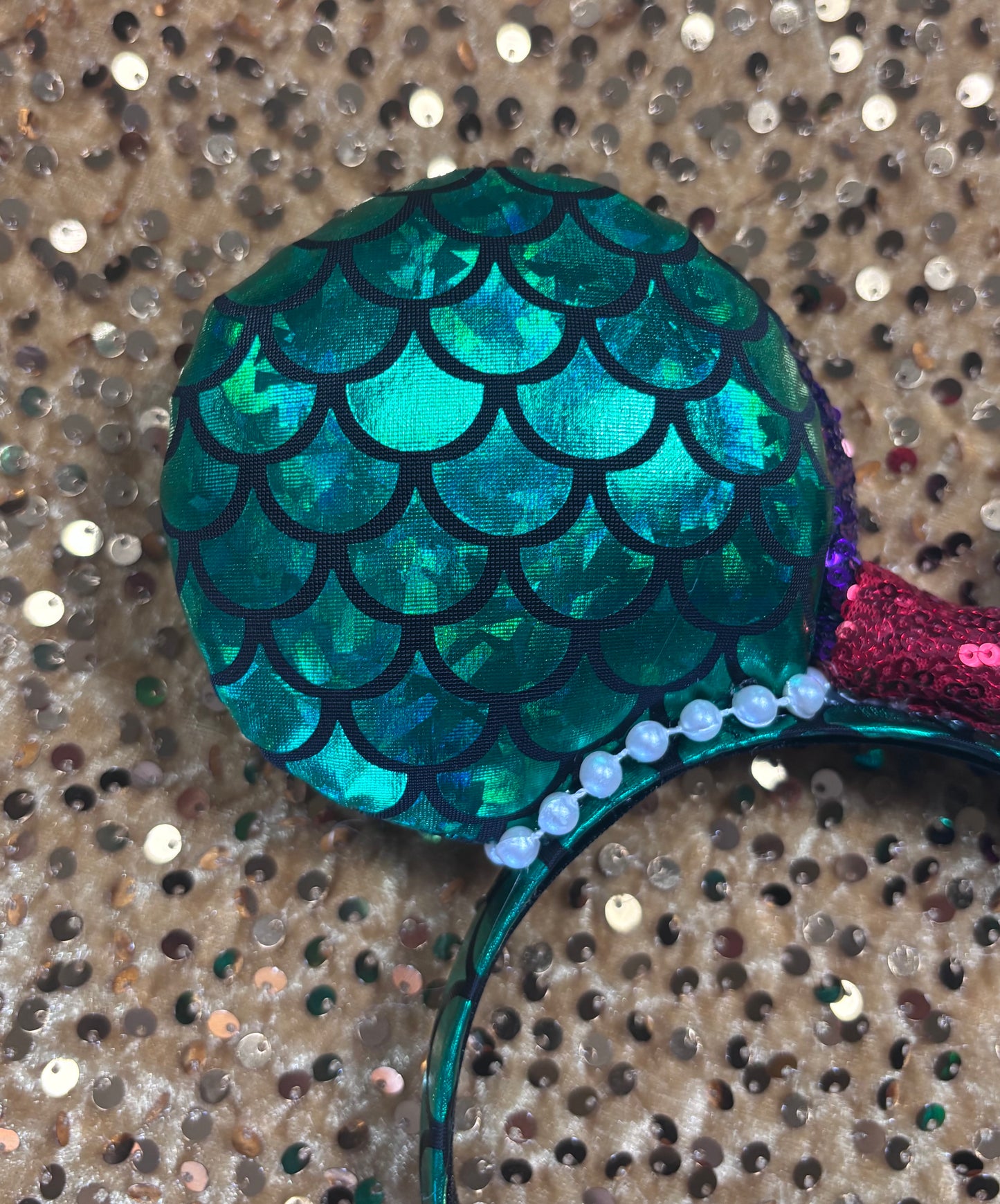 Back ear close up. Mermaid-themed headband with teal scales and a purple sequin bow with a red middle and a dinglehopper middle piece. Pearl garland trim along inner ears m on a matching mermaid scales headband.