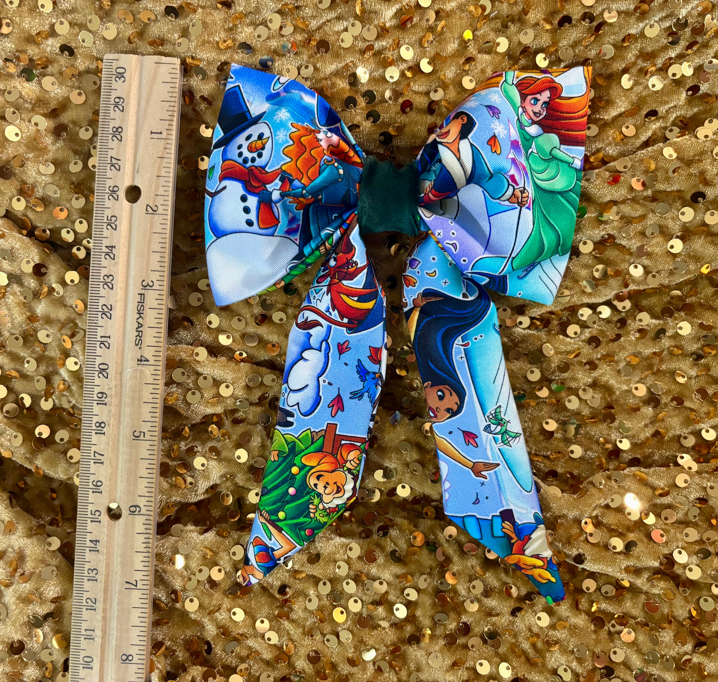 Colorful bow with character designs on a gold sequin background, next to a ruler for scale.