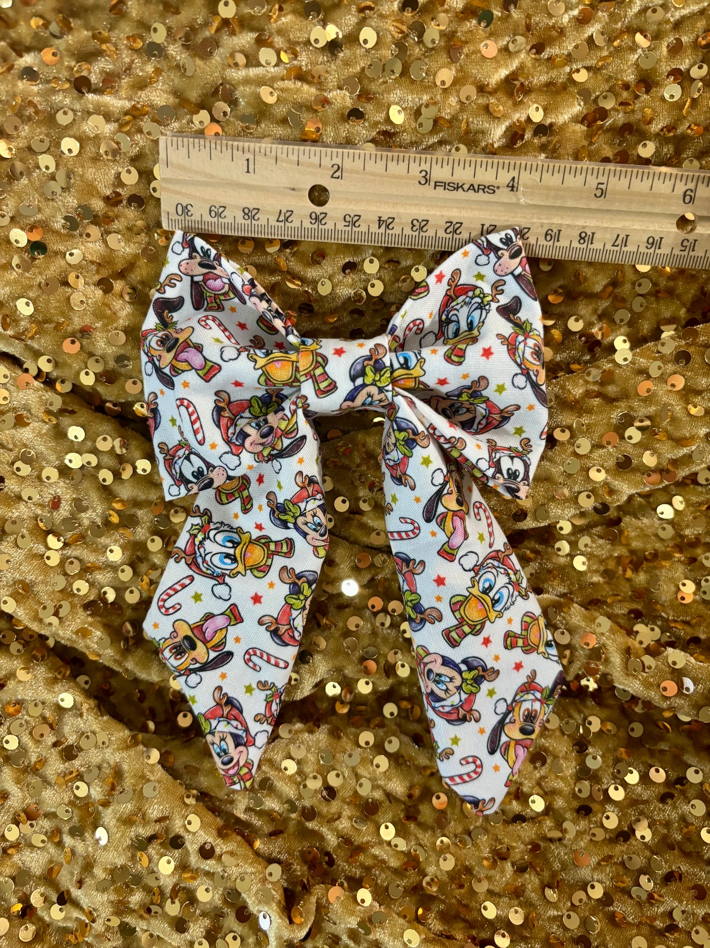 Large fabric bow with cartoon character pattern on a gold sequin background, measured with a ruler.