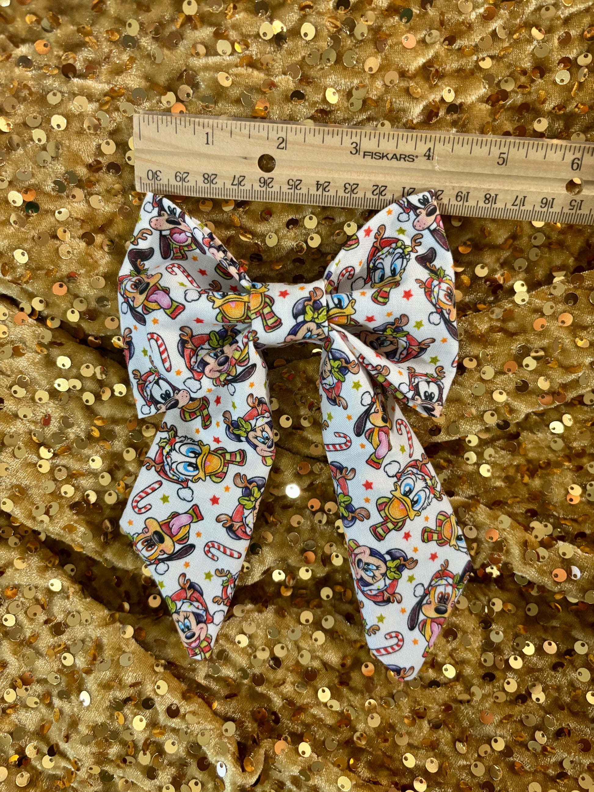 Large fabric bow with cartoon character pattern on a gold sequin background, measured with a ruler.