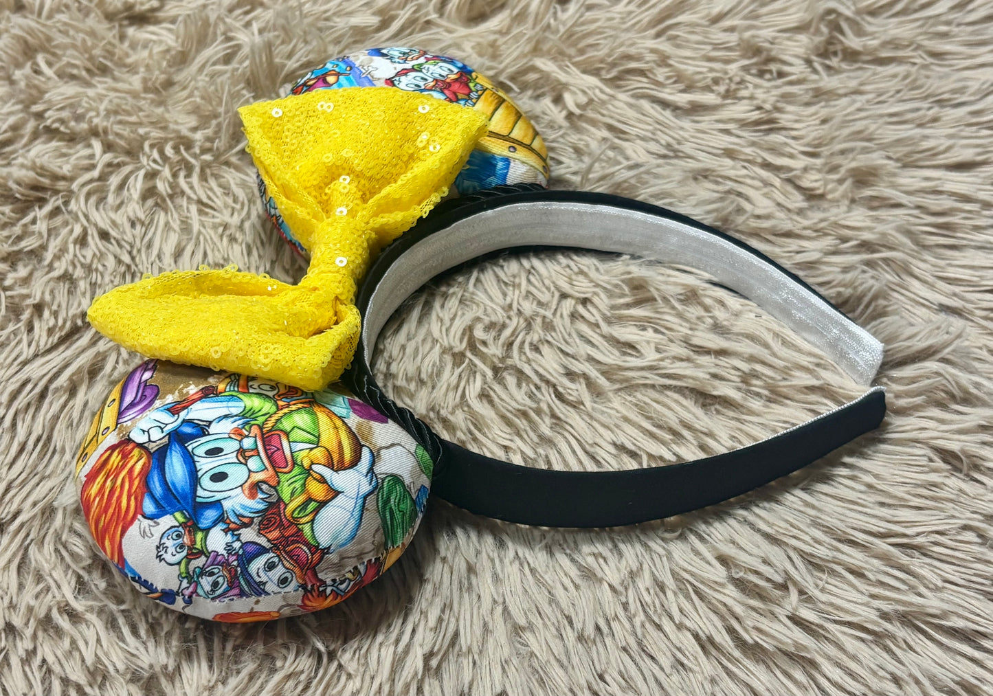 A novelty headband with two round, cartoon-printed ear-like pieces and a large yellow sequined bow, displayed on a beige shag rug.