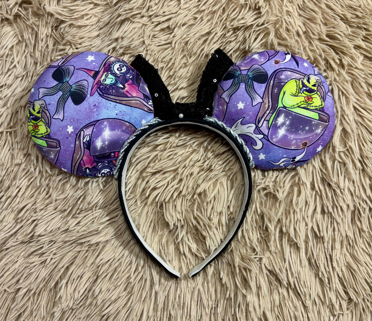 Back side of ears. An ear headband accessory with a black sequined bow, featuring a purple HM and NBC themed print with a cartoon character in a buggy.