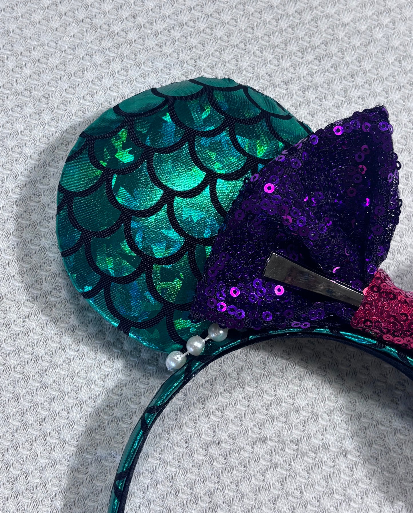 Close up of front ear. Mermaid-themed headband with teal scales and a purple sequin bow with a red middle and a dinglehopper middle piece. Pearl garland trim along inner ears m on a matching mermaid scales headband.