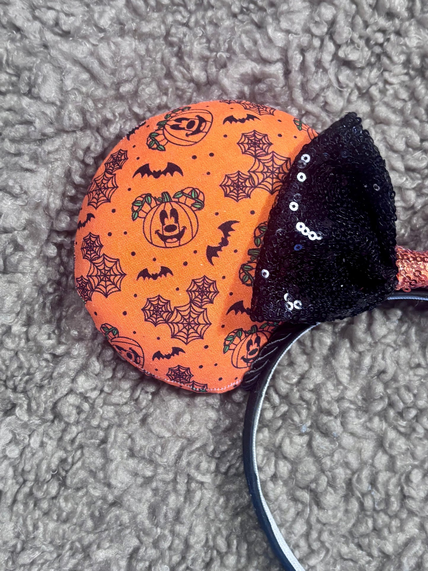 Halloween mouse ears with the fabric on both the front and back. It's orange with bats, mouse shaped pumpkins and spider webs. The headband is black universal sized and fits most. The set has a black with orange middle sequin bow. Item is shown on a brown furry background.