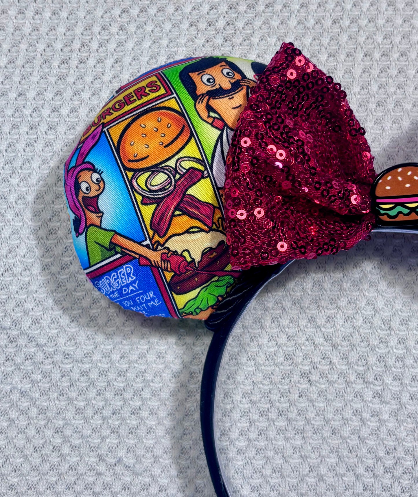 Handmade and ready to ship Belcher and friends mouse ears with the fabric on both the front and back. The headband is black universal sized and fits most. The set has a red sequin bow with a burger embellishment on the middle. Item is shown on a white background.
