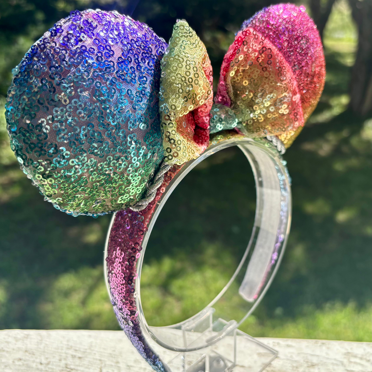 Handmade and ready to ship. Mouse ears with rainbow fabric are on the front and back of mouse ears along with the headband with a matching rainbow sequin bow. Shown on a white background.