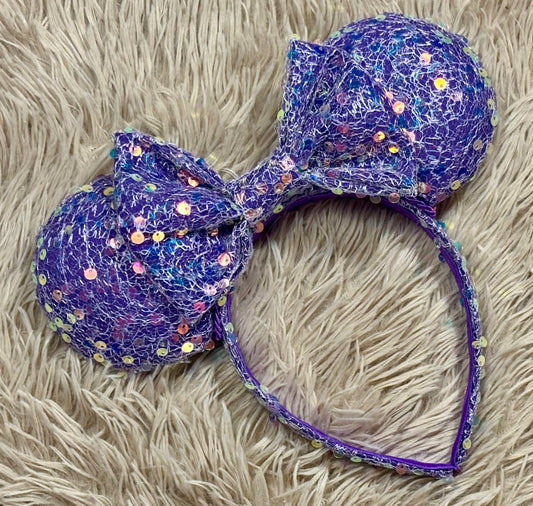 A purple sequined headband with large round ears and a bow, laid flat on a beige faux fur surface.