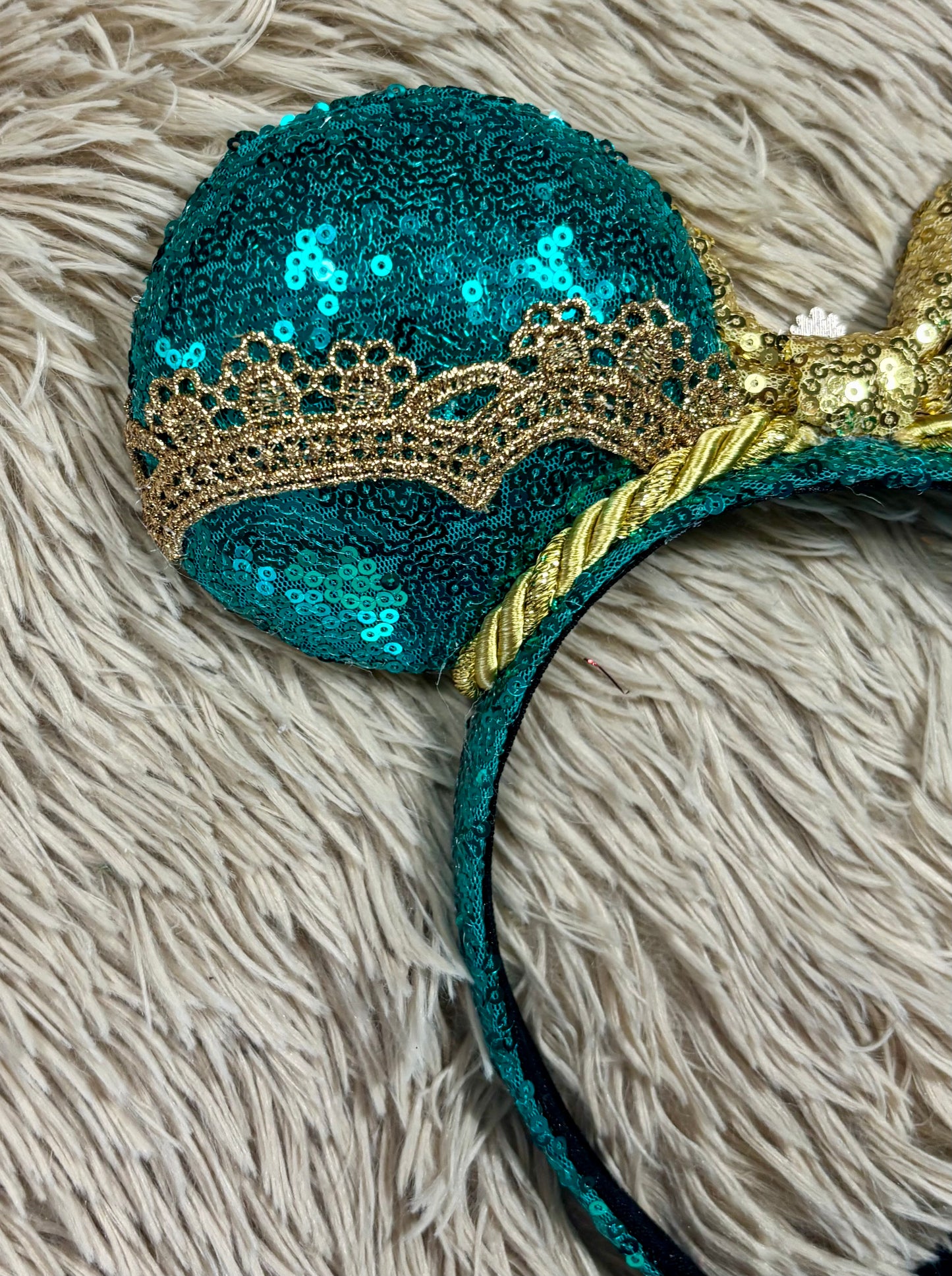 A close-up of a teal sequined headband with a large round ear-shaped section, a gold lace crown detail, and a gold rope accent on a textured beige faux fur background.