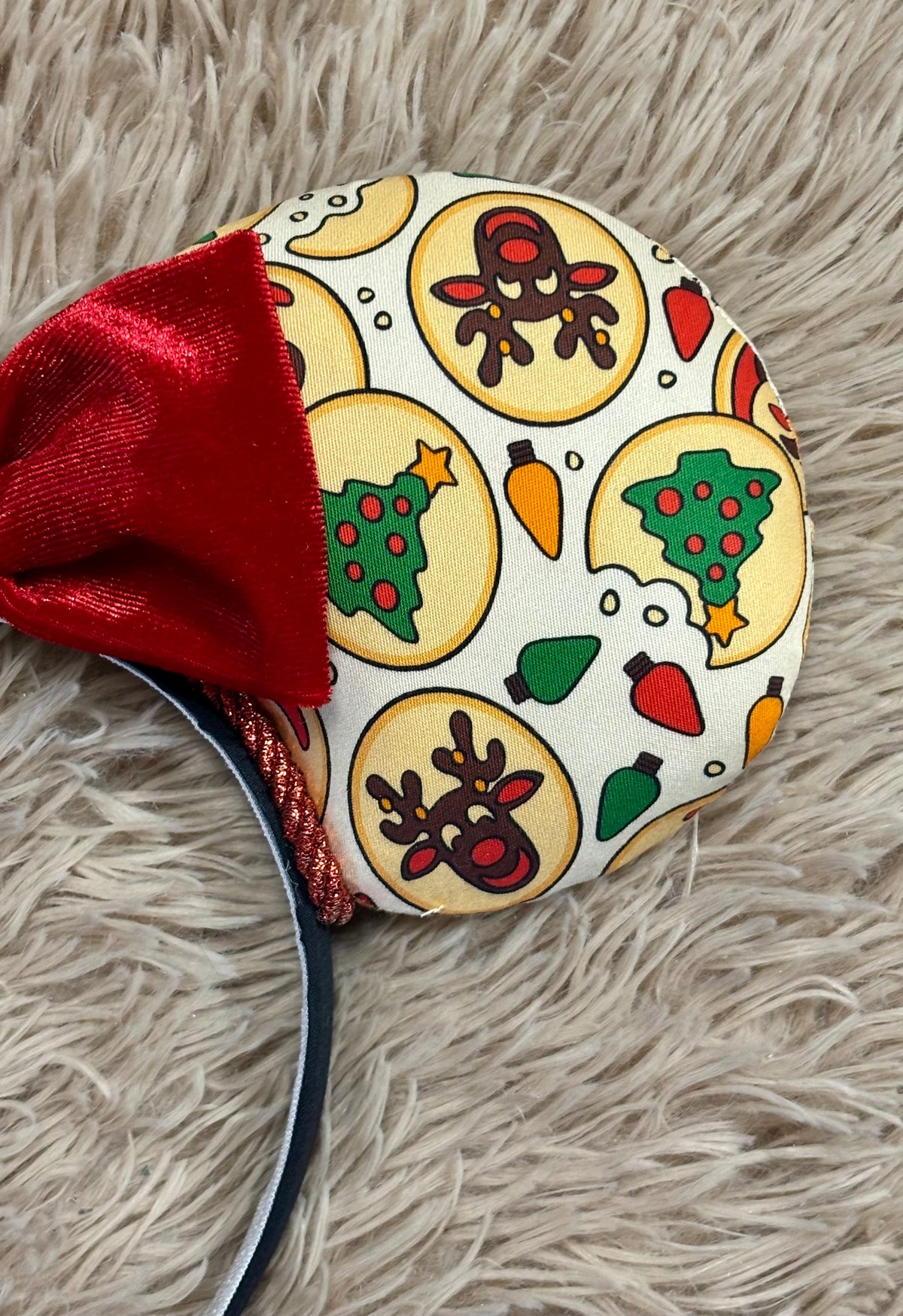 An ear headband featuring a festive Christmas print with red velvet bows, displayed on a beige faux fur background.