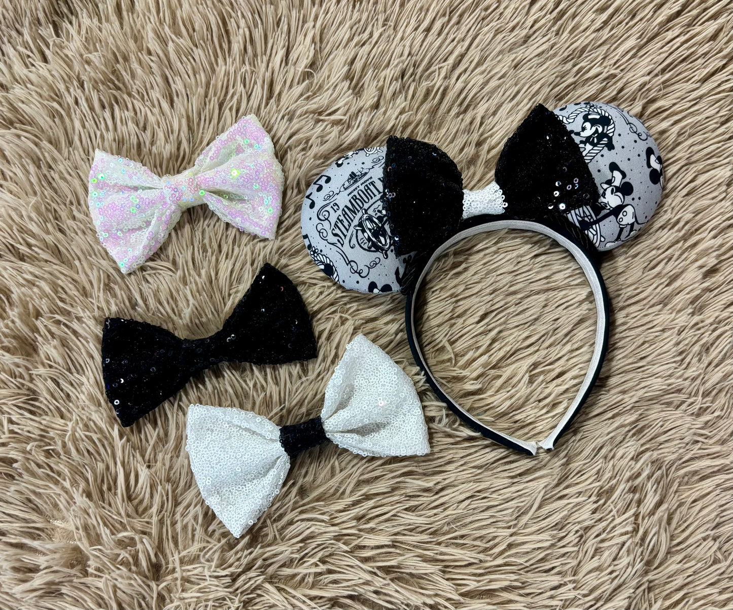 A pair of Mouse-themed ear headbands featuring a black and white patterned fabric with sequins, a black sequined bow with a white sequin middle, and a white headband, displayed on a beige faux fur background.