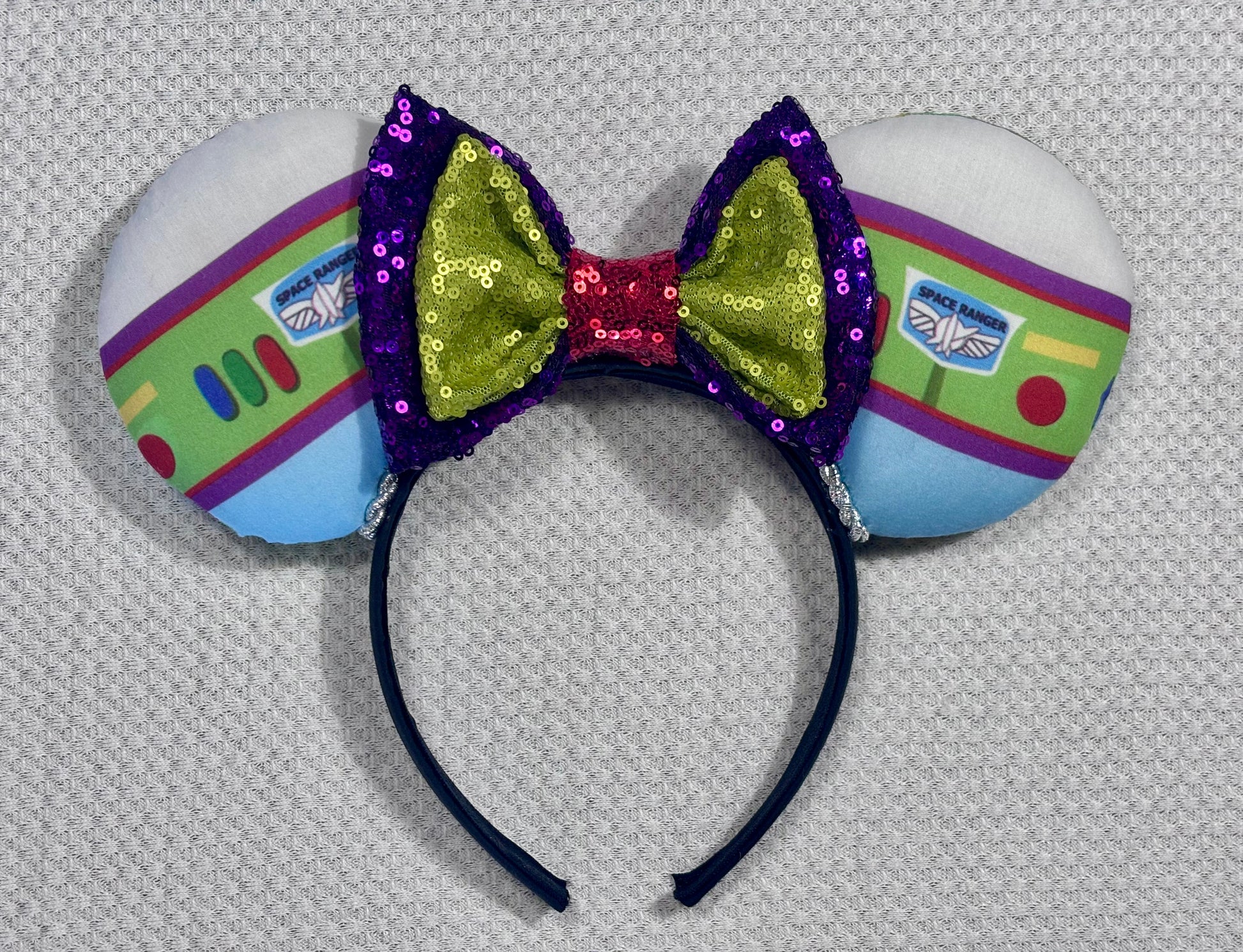 Space Ranger Mouse Ear headband with new improved bow. Handmade and ready to ship with Space ranger fabric on both the front and the back of mouse ears. The set has a silver twisted trim along the inner ears and a new sequin bow that is purple, lime green and red in sequin. Ears are on a black poly cotton universal sized headband.