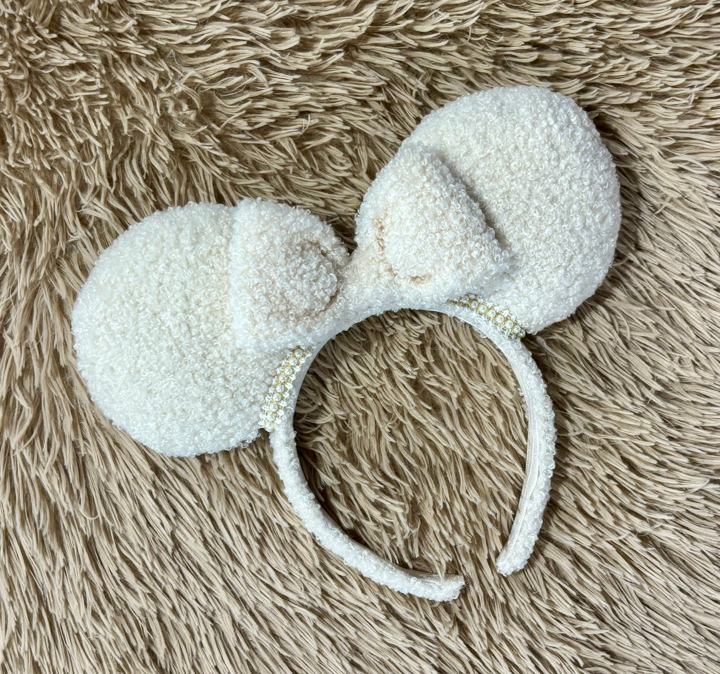 A pair of fluffy cream-colored ears on a headband with a matching bow on a textured beige shag rug.