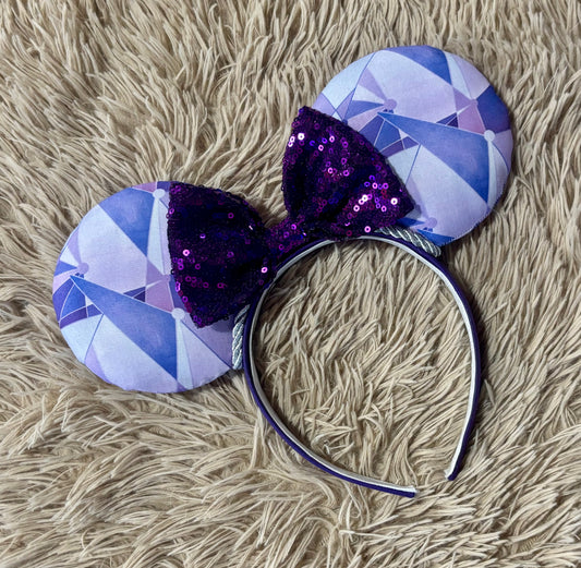 A headband with round ears. The ears have a geometric pattern in shades of purple, and white, while the bow on top is made of dark purple sequins.